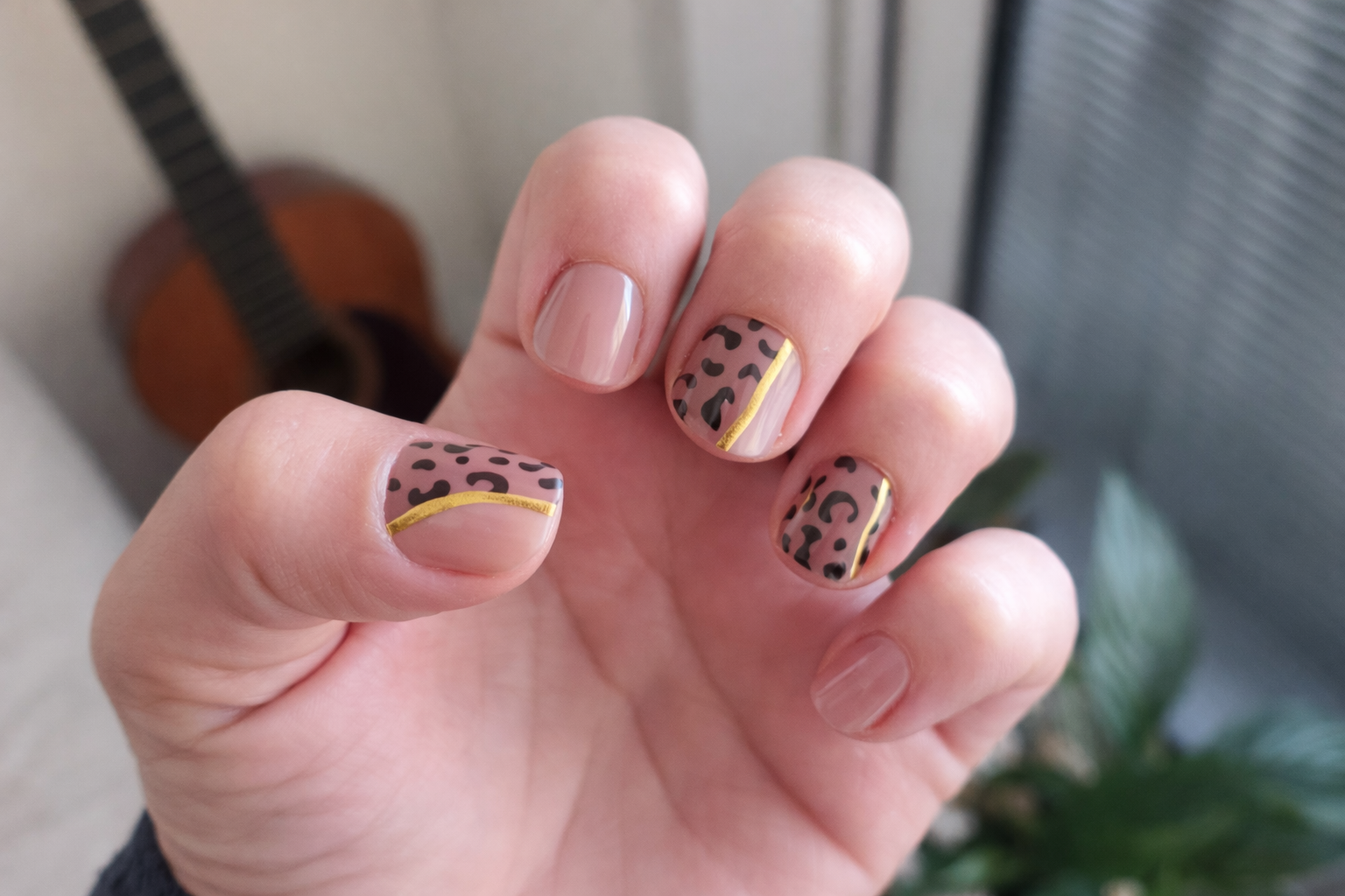 Hand with leopard print nail design holding a small object, with a guitar and plant in the background.